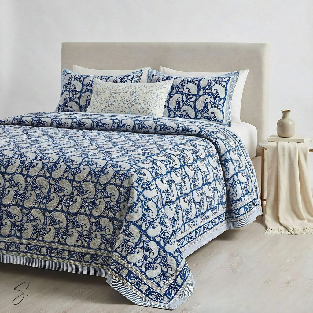 A neatly made bed with a blue and cream paisley-patterned quilt, matching pillow shams, and a small decorative accent pillow, styled in a minimalist bedroom setting with a beige headboard, side table, and neutral decor.