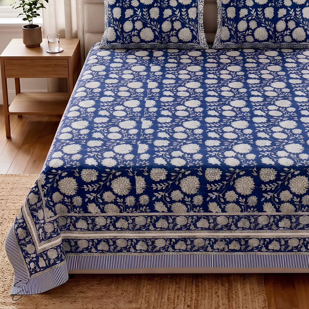 A bed dressed in a navy blue and off-white floral block-print style bedspread and matching pillow shams, shown in a styled bedroom setting with a wooden side table, plant, and woven rug.