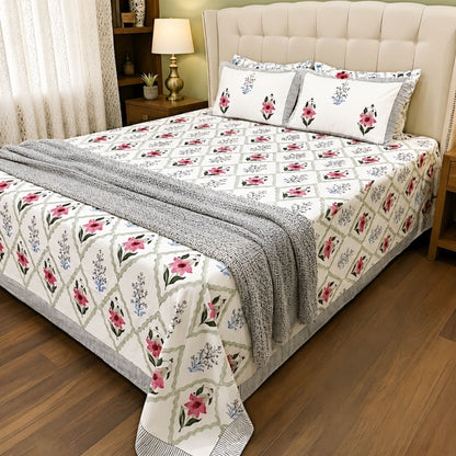 A neatly made bed in a bedroom setting featuring a cream-colored quilted tufted headboard, a patterned quilt with a diamond lattice design, pink and blue floral motifs, matching floral pillows, a gray knit throw blanket, and a brown wood floor.