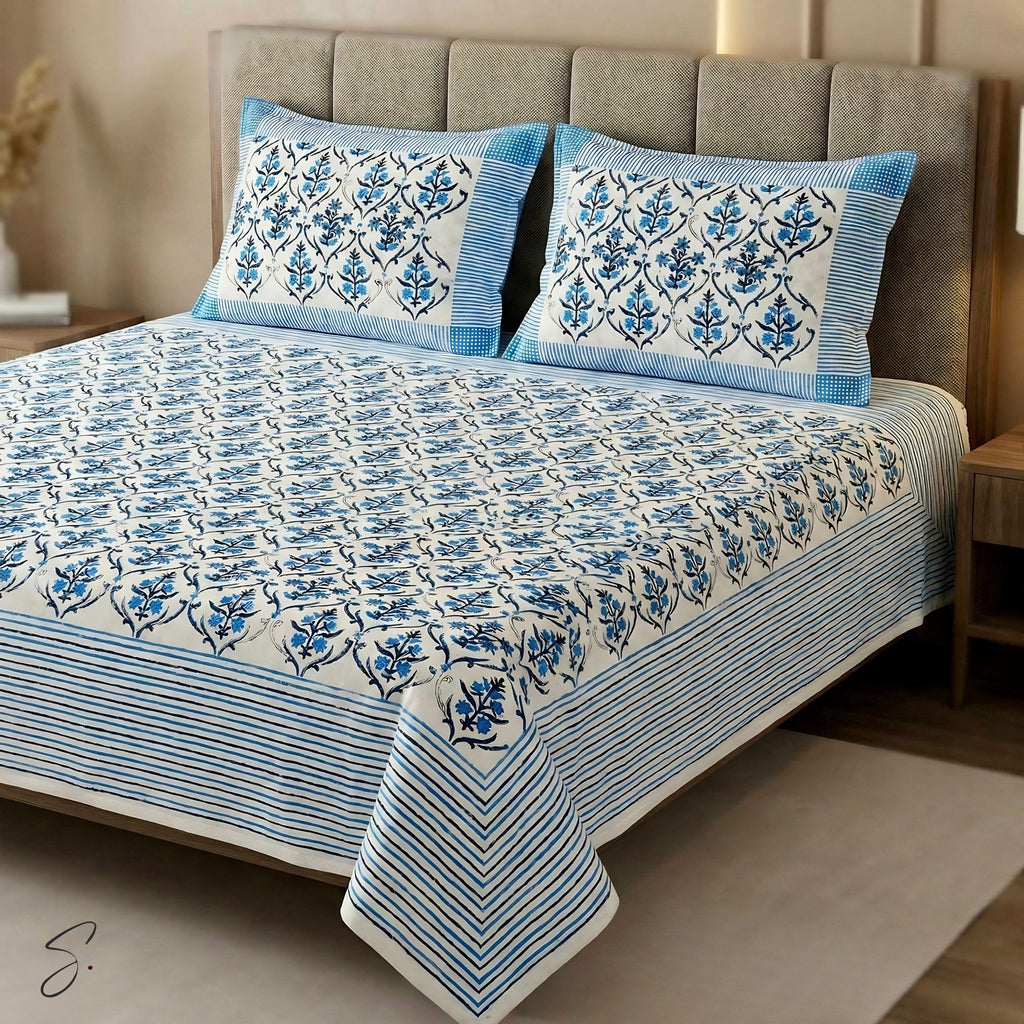 A blue and white block-print cotton bedding set, including a bedspread and two matching pillowcases, styled on a bed with a gray upholstered headboard in a bedroom setting.