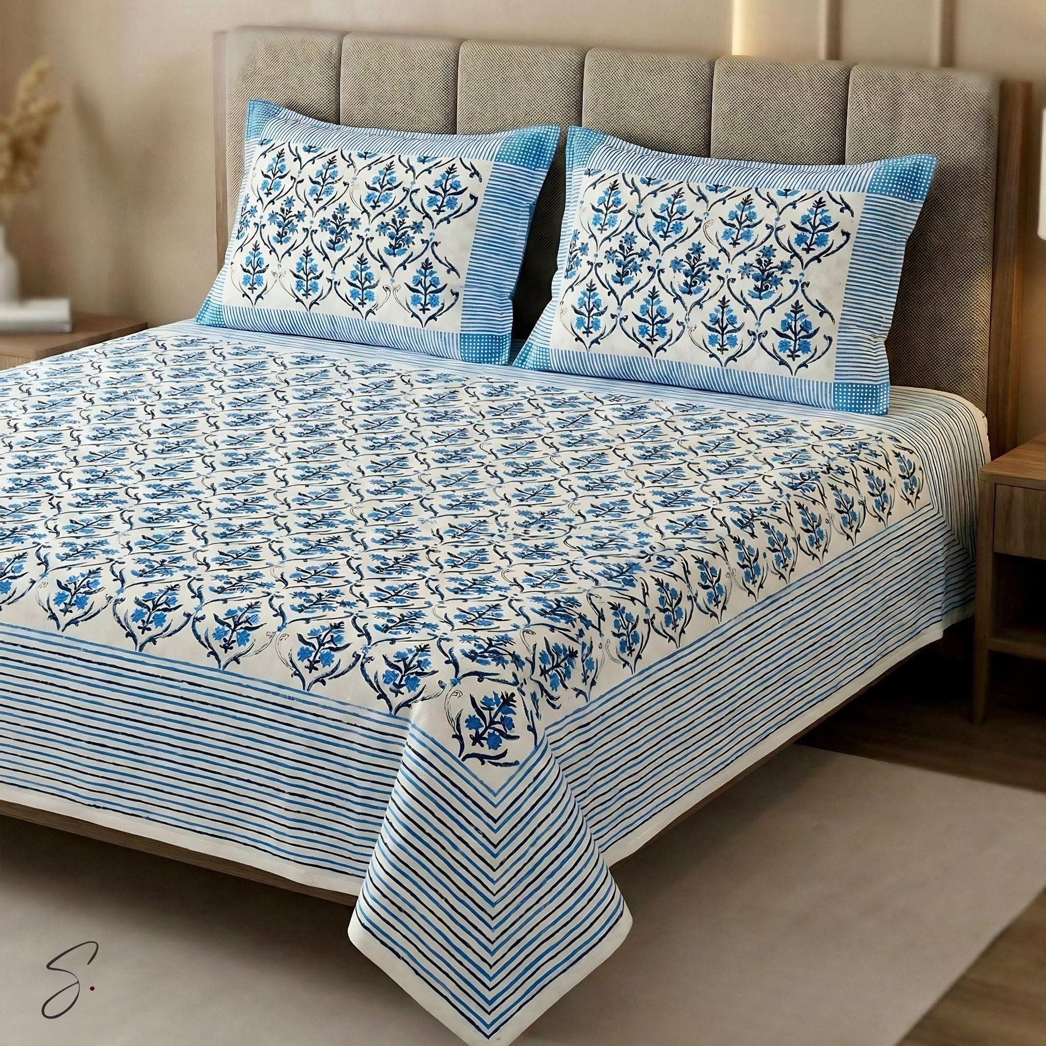A blue and white block-print cotton bedding set, including a bedspread and two matching pillowcases, styled on a bed with a gray upholstered headboard in a bedroom setting.