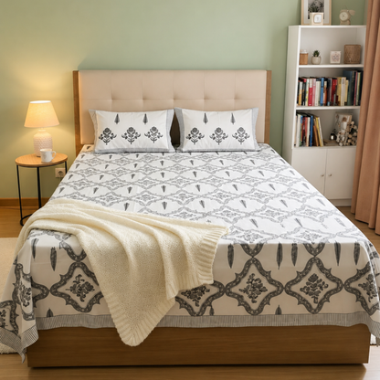 A neatly made bed in a styled bedroom, featuring a white bedding set with a gray geometric damask pattern, a beige tufted headboard, a cream knit throw blanket draped over the foot of the bed, a lit table lamp on the left, and a white bookshelf on the right.
